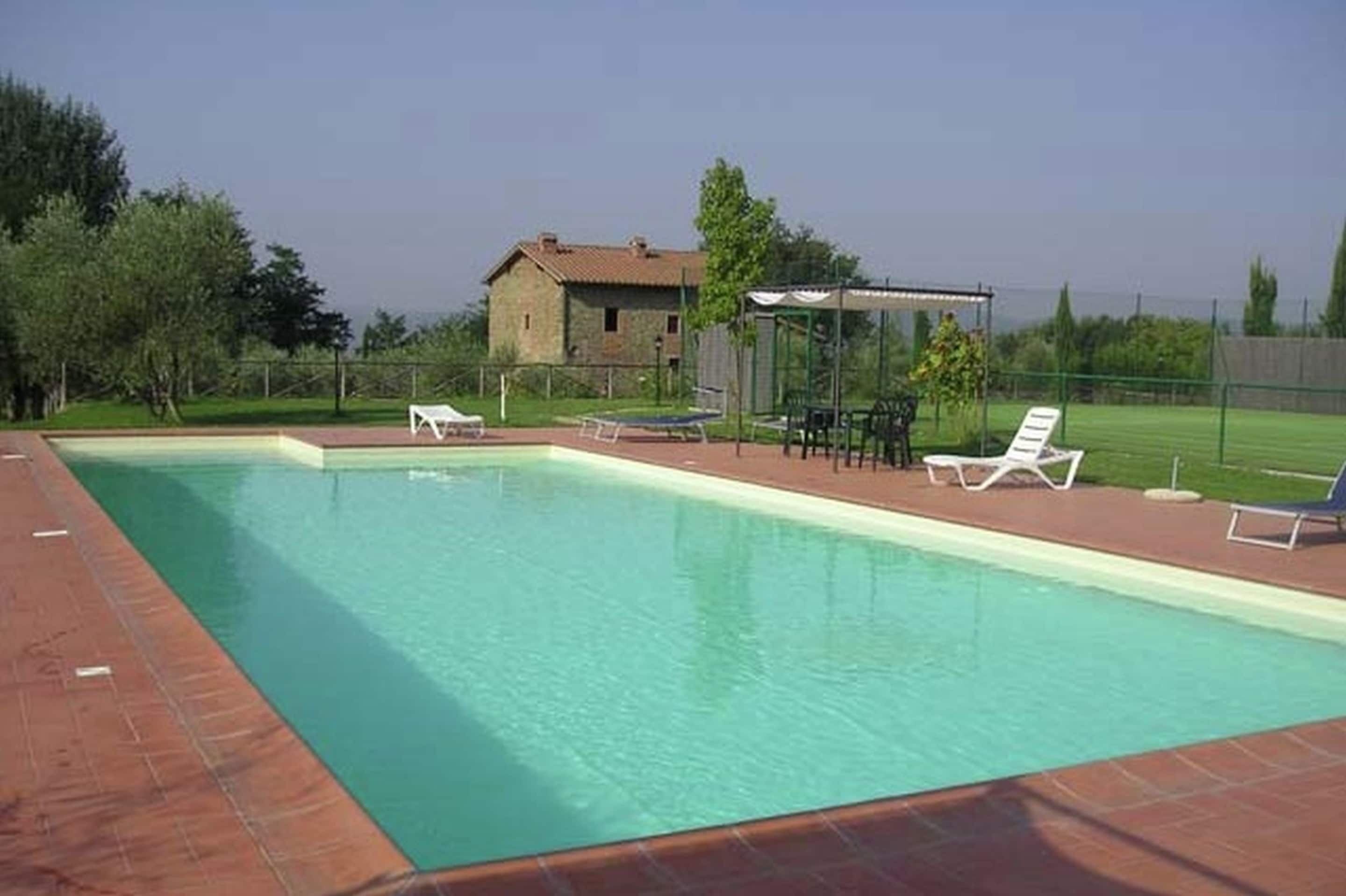 A rectangular swimming pool with a tiled edge, surrounded by a brick deck. There are lounge chairs and a covered seating area with a table and chairs. A small building with a sloped roof is visible in the background.