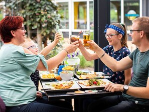A group of people toasting with drinks at a table with various dishes, including pizza and salad, in an outdoor setting with greenery in the background.