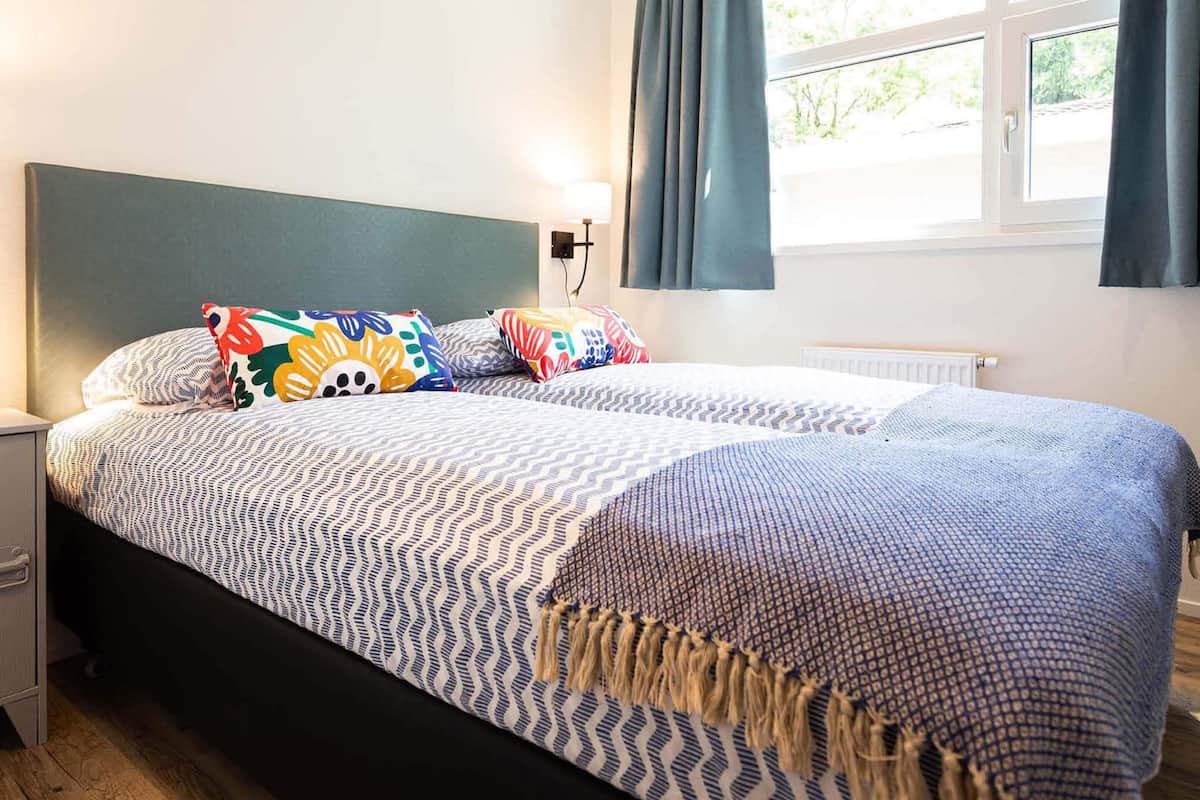 A neatly made bed with patterned bedding, a bedside table, a wall-mounted lamp, and a window with curtains.