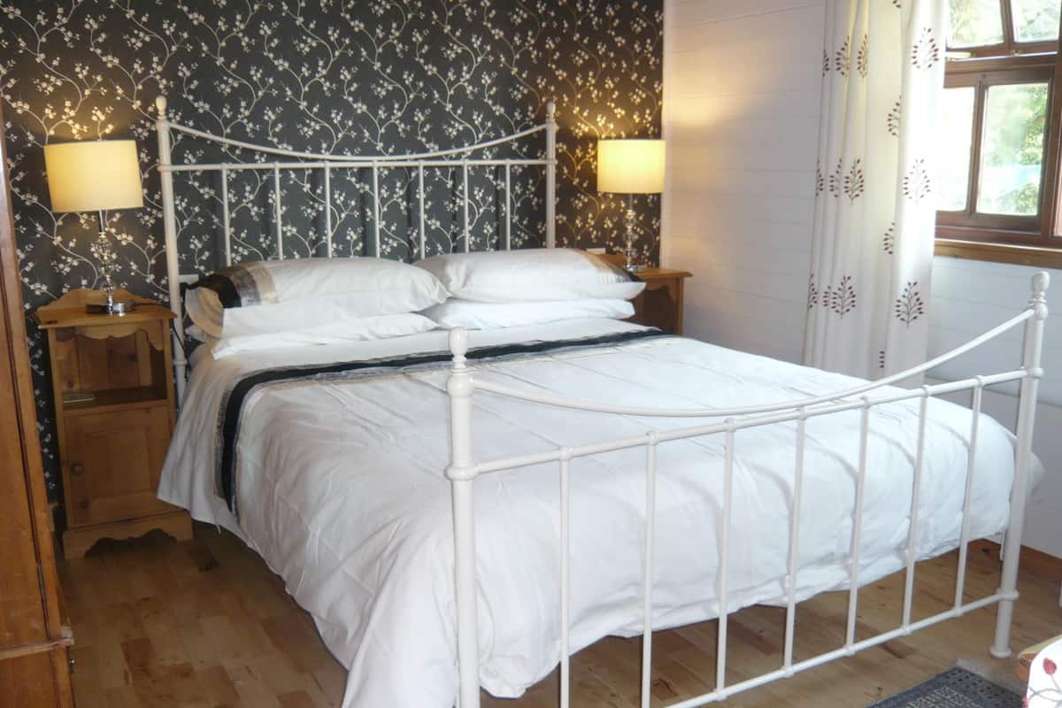 Una cama con estructura metálica blanca, dos mesitas de noche, un buró, un papel tapiz floral y una ventana con cortinas.