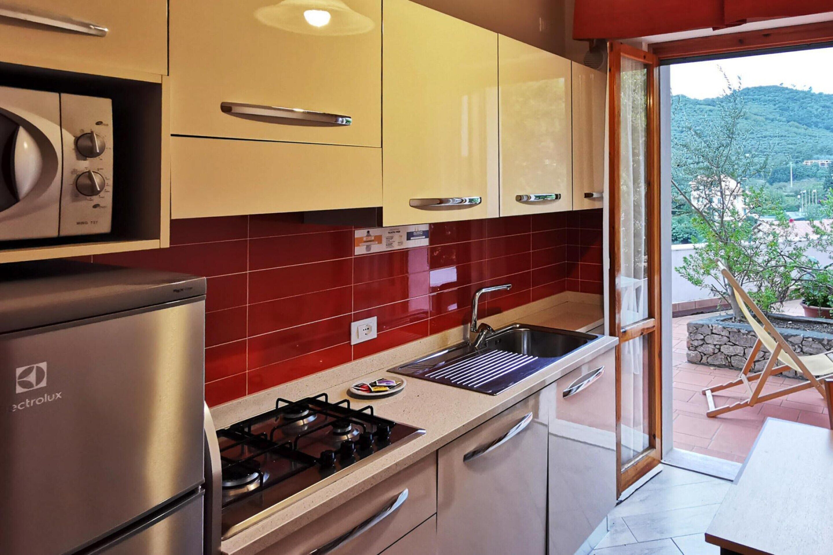 Une cuisine équipée d’une cuisinière à gaz, d’un four à micro-ondes et d’un évier. On peut admirer, depuis la cuisine, un balcon avec une chaise et un pot de fleurs.
