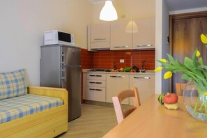 Une cuisine compacte équipée d’un réfrigérateur, d’un four à micro-ondes et d’une cuisinière. On y trouve une table à manger avec des chaises et un petit canapé. Un vase rempli de fleurs est posé sur la table.