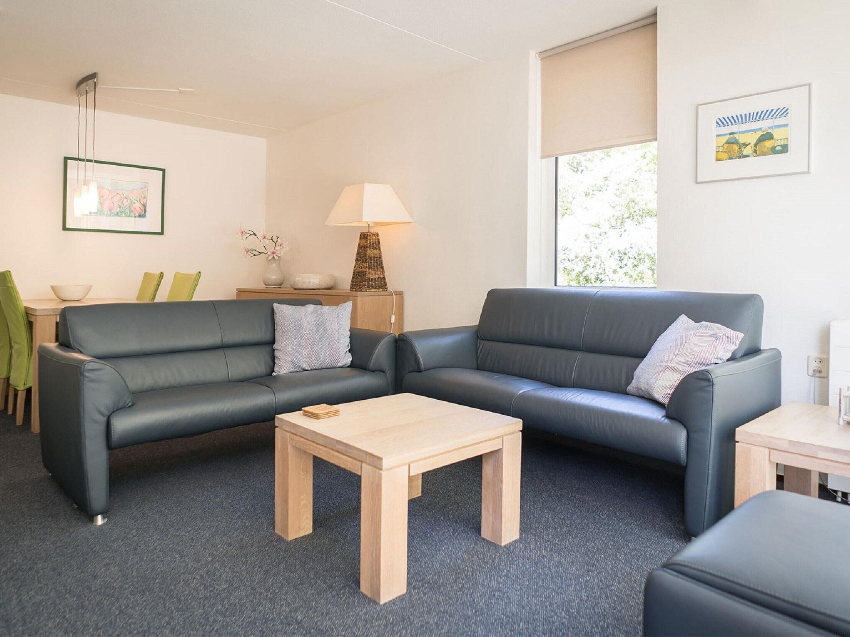 Ein modernes Wohnzimmer mit grauem Ledersofa, einem Holztisch und einem Essbereich mit einer Schüssel und grünen Stühlen.