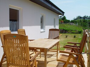 Eine Holzterrasse mit Tisch und Stühlen, ein weißes Gebäude mit Fenstern und ein Garten mit Gras und Pflanzen.