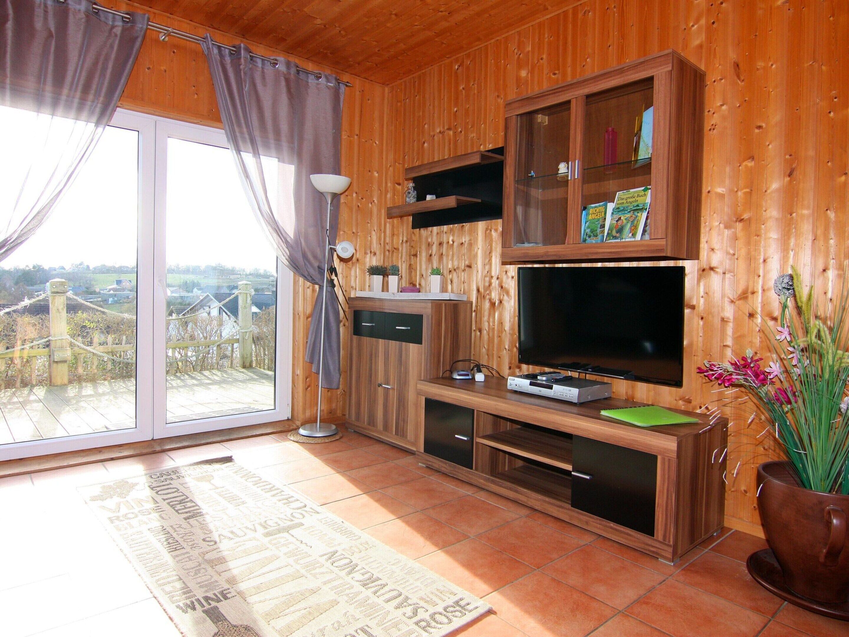 A wooden-paneled room with a large window, a flat-screen TV, a bookshelf, and a potted plant.