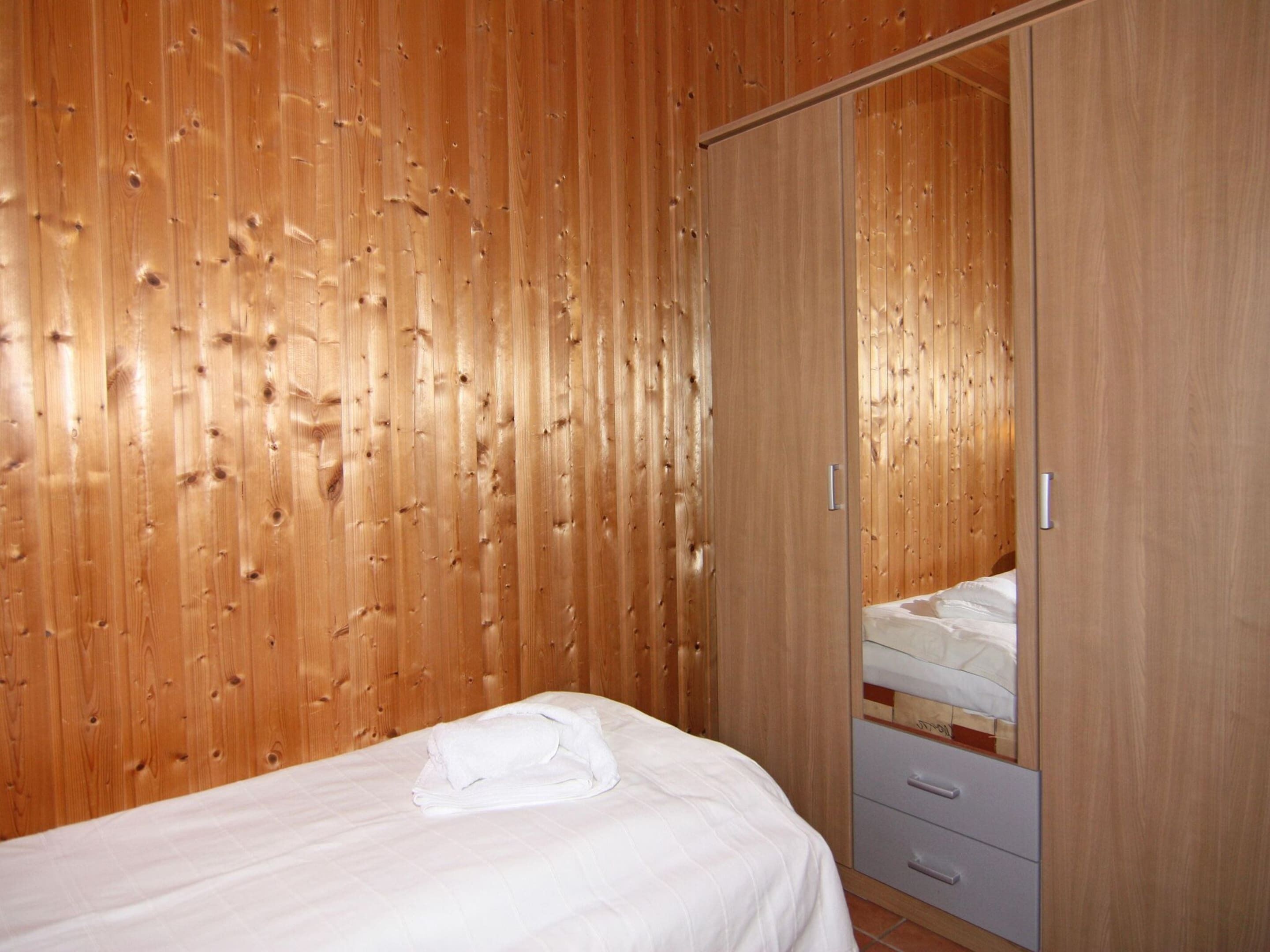 A bedroom with a wooden wall, a bed with a white duvet, and a wardrobe with a mirror.