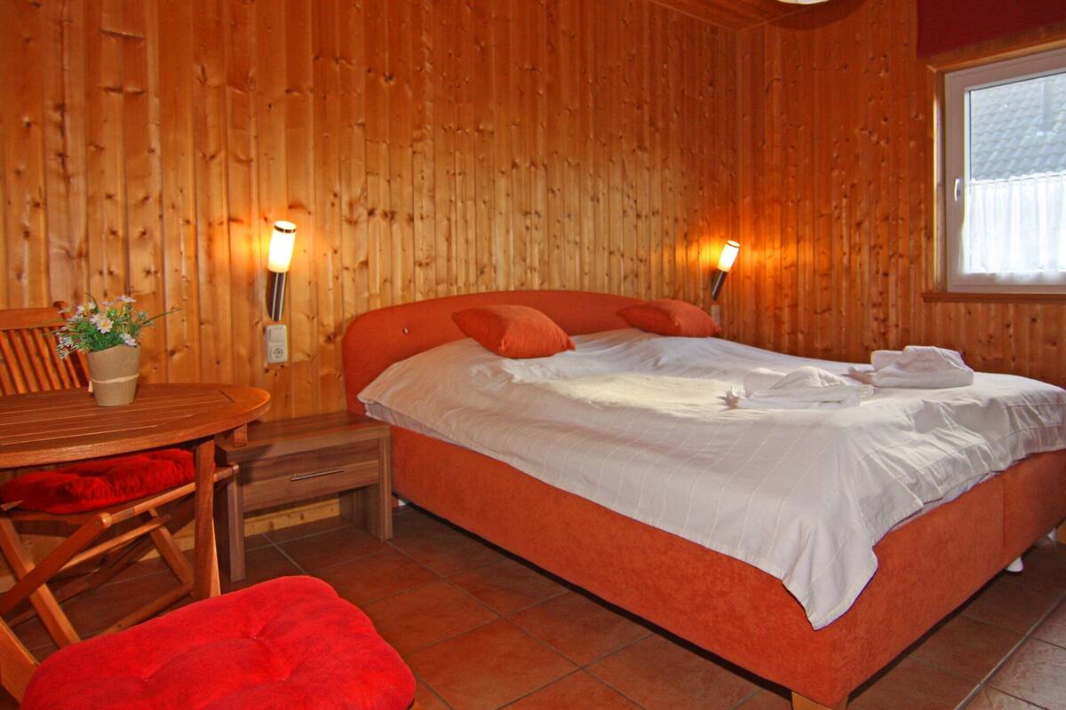 A wooden room with a bed, a table with a plant, and a window.