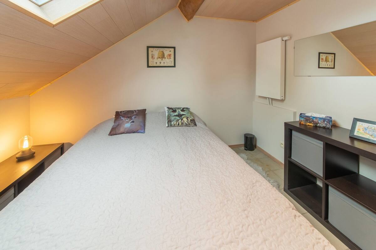 A bedroom with a sloped ceiling, a bed with two pillows, a bedside table with a lamp, a trash can, and a dresser with a mirror and a framed picture.
