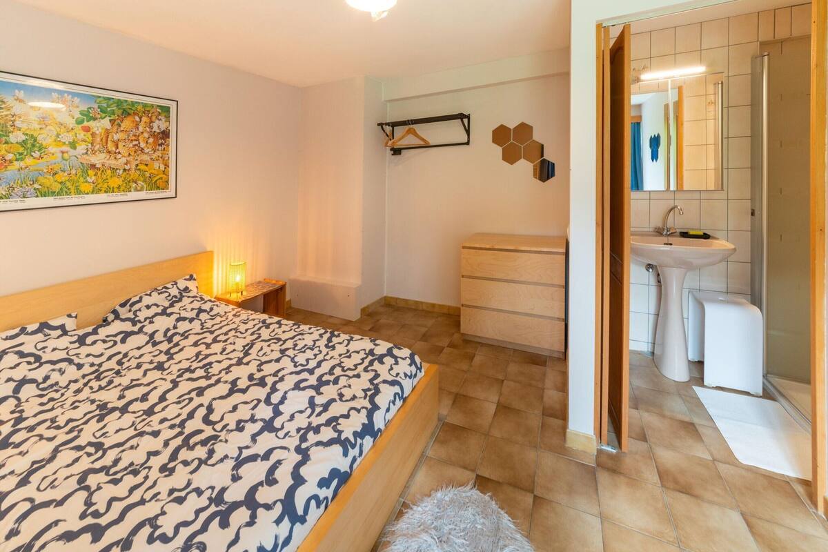 A bedroom with a patterned bedspread, a bedside table with a lamp, a dresser, and a wall-mounted shelf.