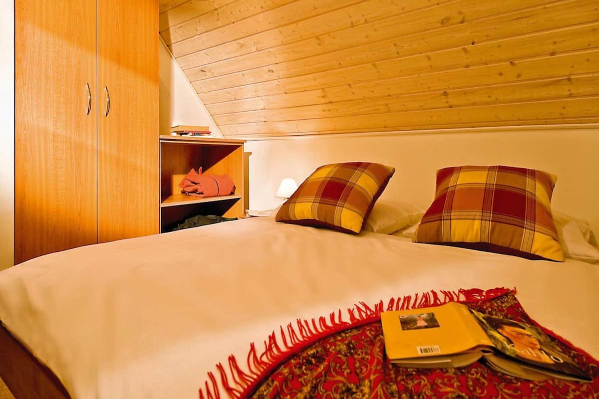 A bedroom with a wooden ceiling, a bed with a red and yellow patterned blanket, two pillows, a nightstand with a lamp, and a wardrobe.