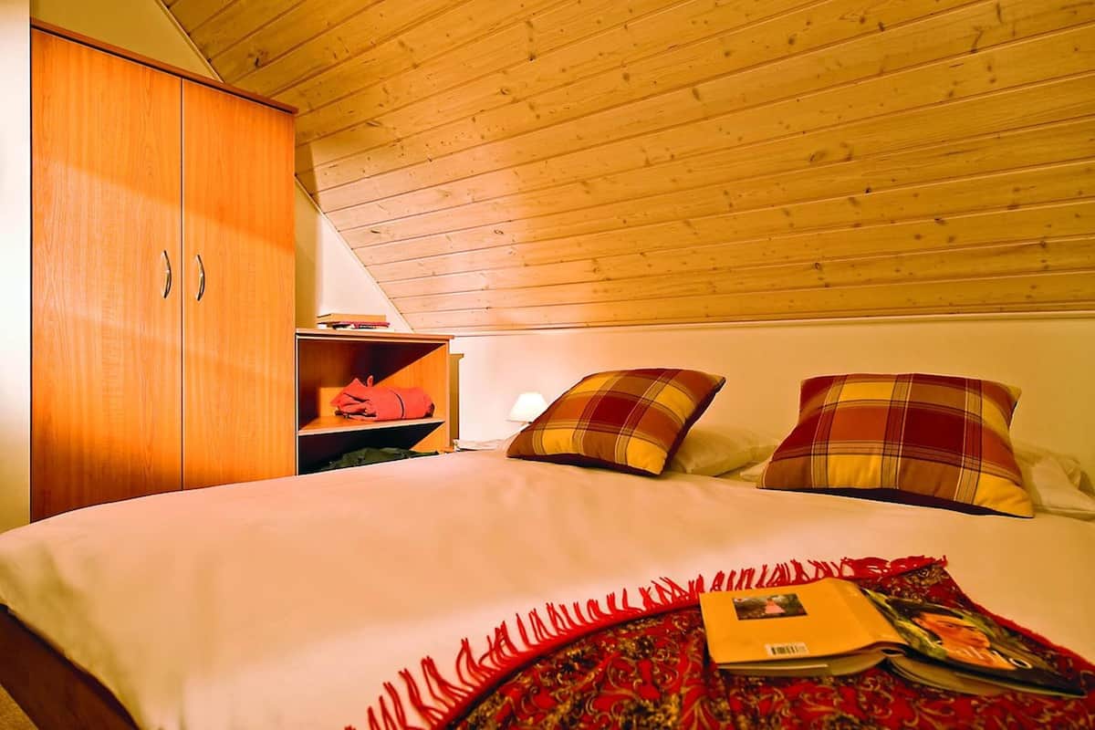 A bedroom with a wooden ceiling, a bed with plaid pillows, a red blanket, and a shelf with a red bag.