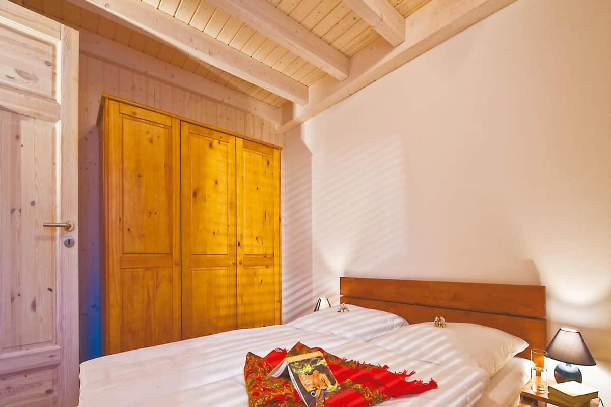 A bedroom with a wooden headboard, a large bed with white bedding, a red decorative pillow, and a nightstand with a lamp and a glass of water.