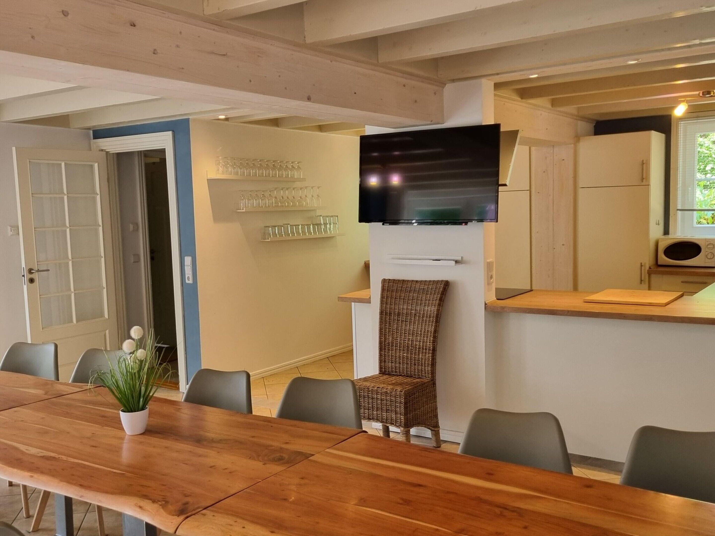 A dining area with a long wooden table, chairs, a television mounted on the wall, and a kitchen area with a microwave and cabinets.