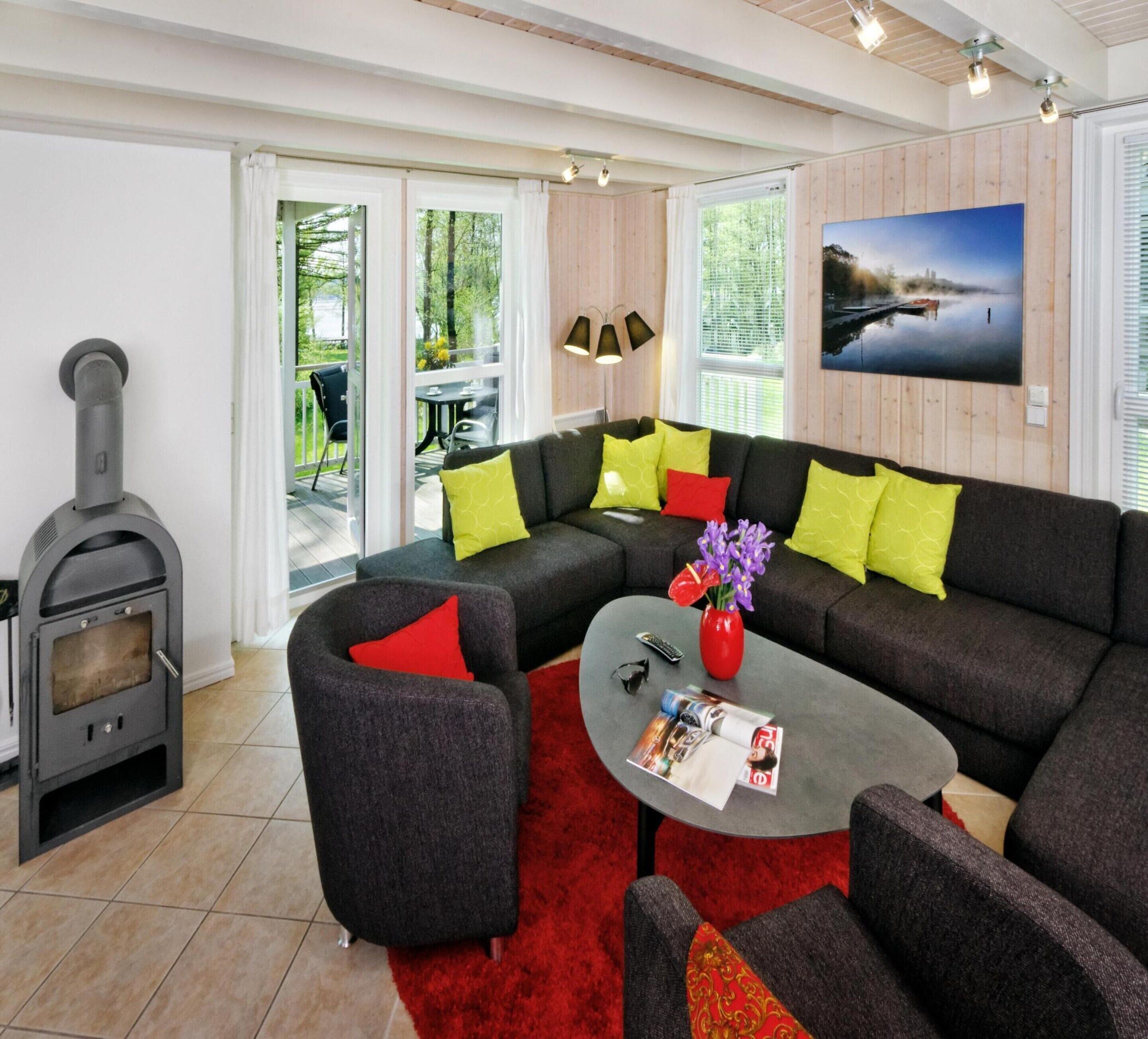 A modern living room with a sofa set, a red rug, a coffee table with magazines, a vase with flowers, and a wood-burning stove.