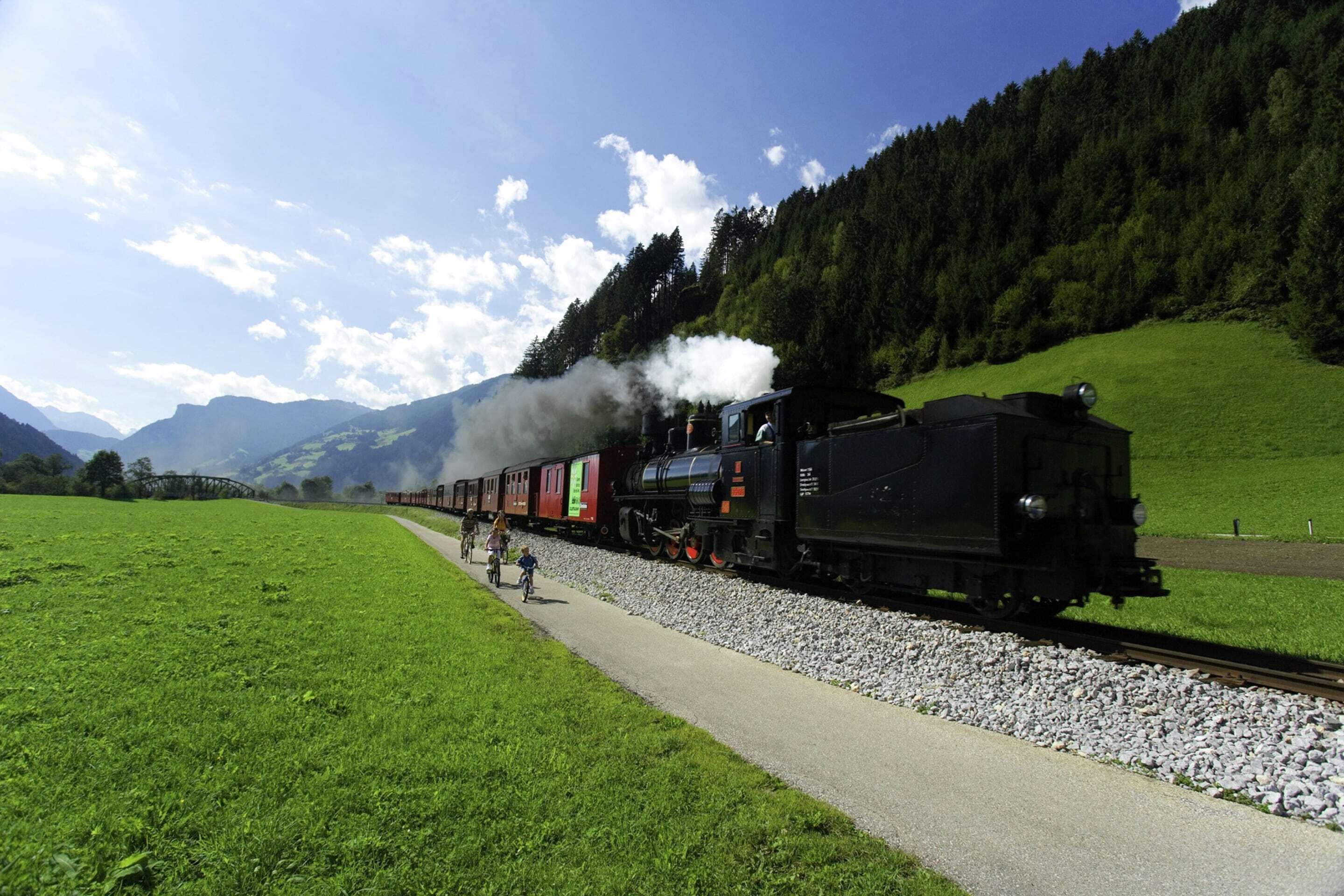 Un train transportant des passagers et des cyclistes circule sur une voie ferrée bordée d’un champ verdoyant et de collines boisées.