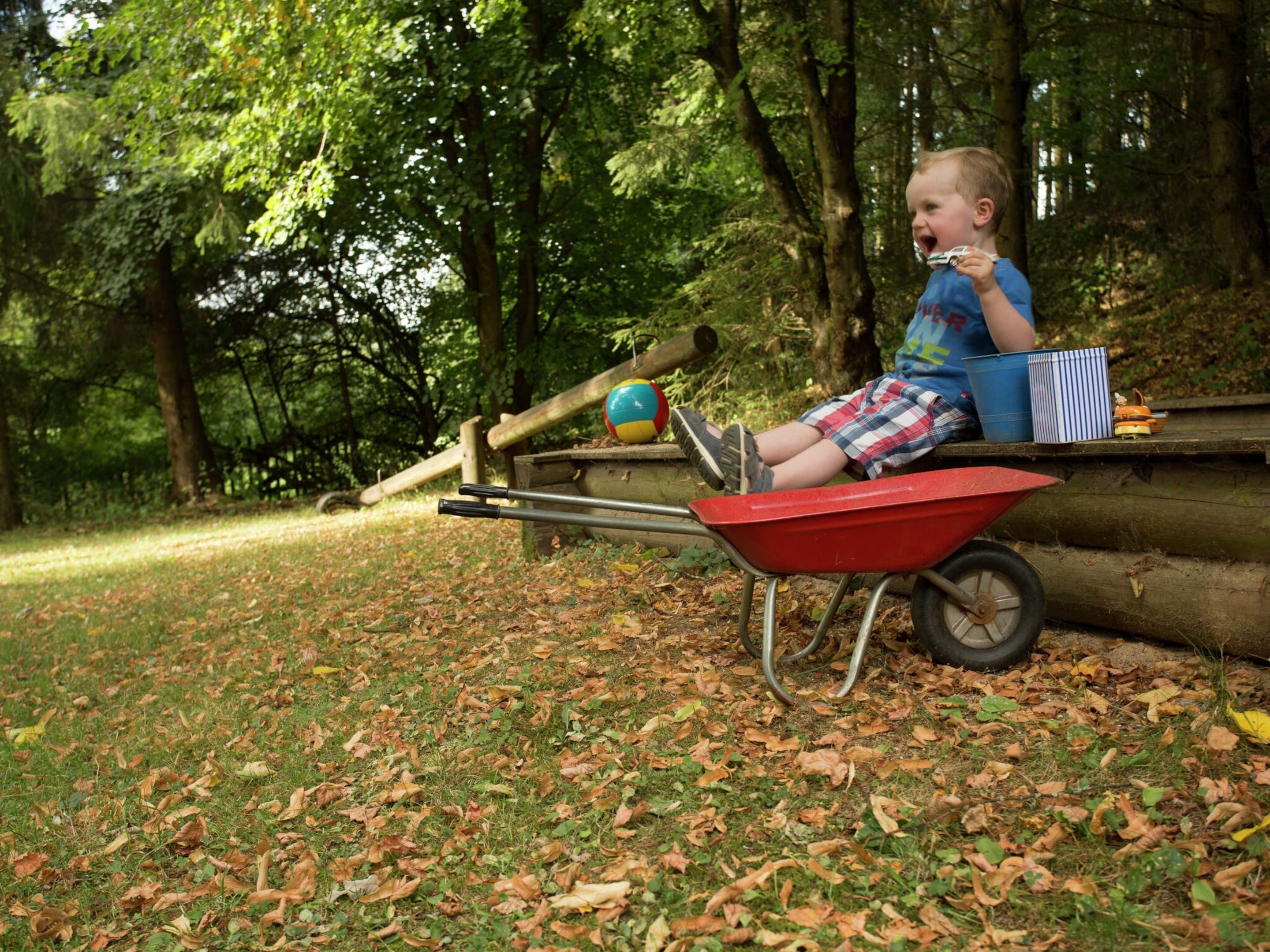Un enfant est assis sur une brouette rouge dans un parc, avec des arbres et une aire de jeux en arrière-plan.