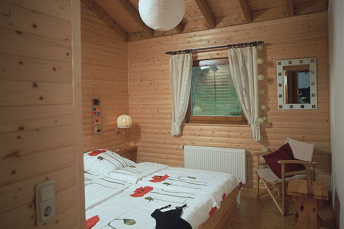 A wooden cabin bedroom with a bed, a chair, a window with curtains, a mirror, and a radiator.