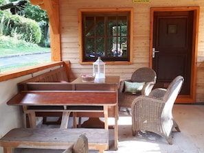 A wooden cabin with a bench and wicker chairs, a table with a lantern, and a door with a sign.