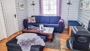 A cozy living room with a blue sofa, a wooden coffee table, a fluffy ottoman, a vintage stove, and a window with purple curtains. - 8 Person Holiday Home in Aseral (Aseral)