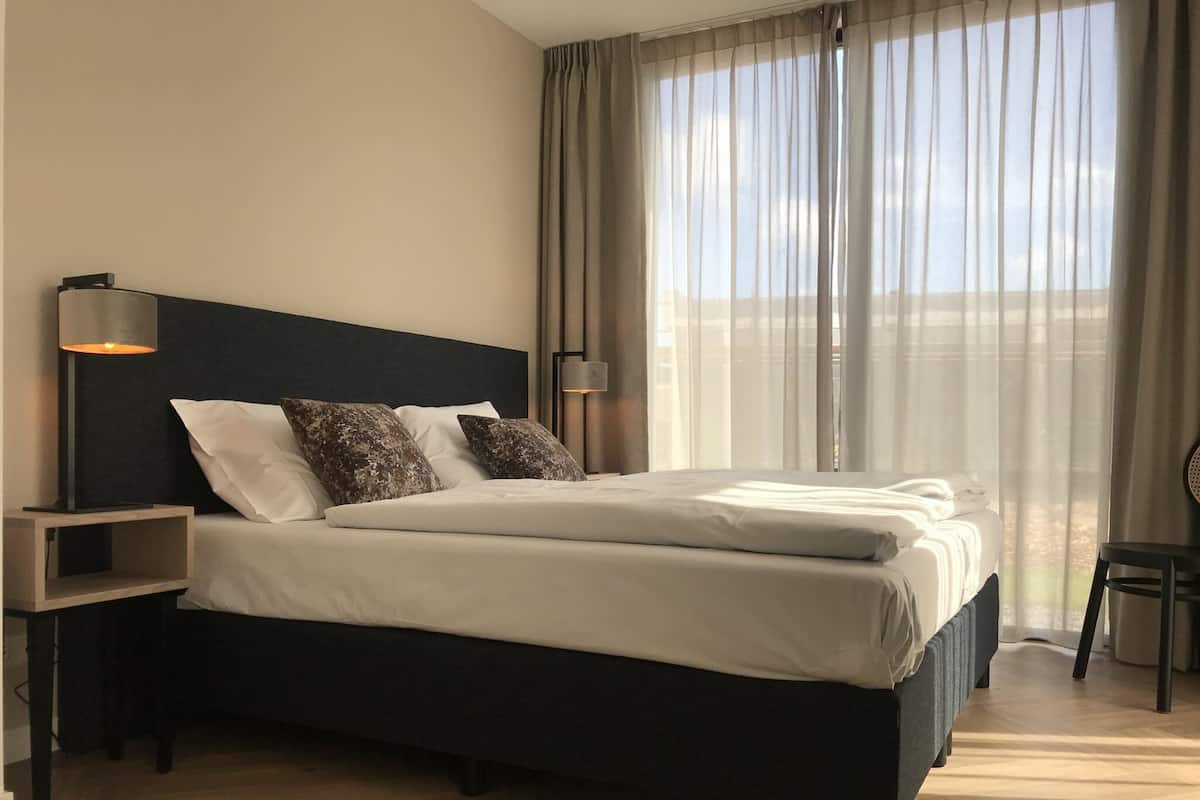 Moderni hotellihuone, jossa on suuri sänky, kaksi yöpöytävalaisinta, yöpöytä ja näkymä ulos ohuiden verhojen läpi.
