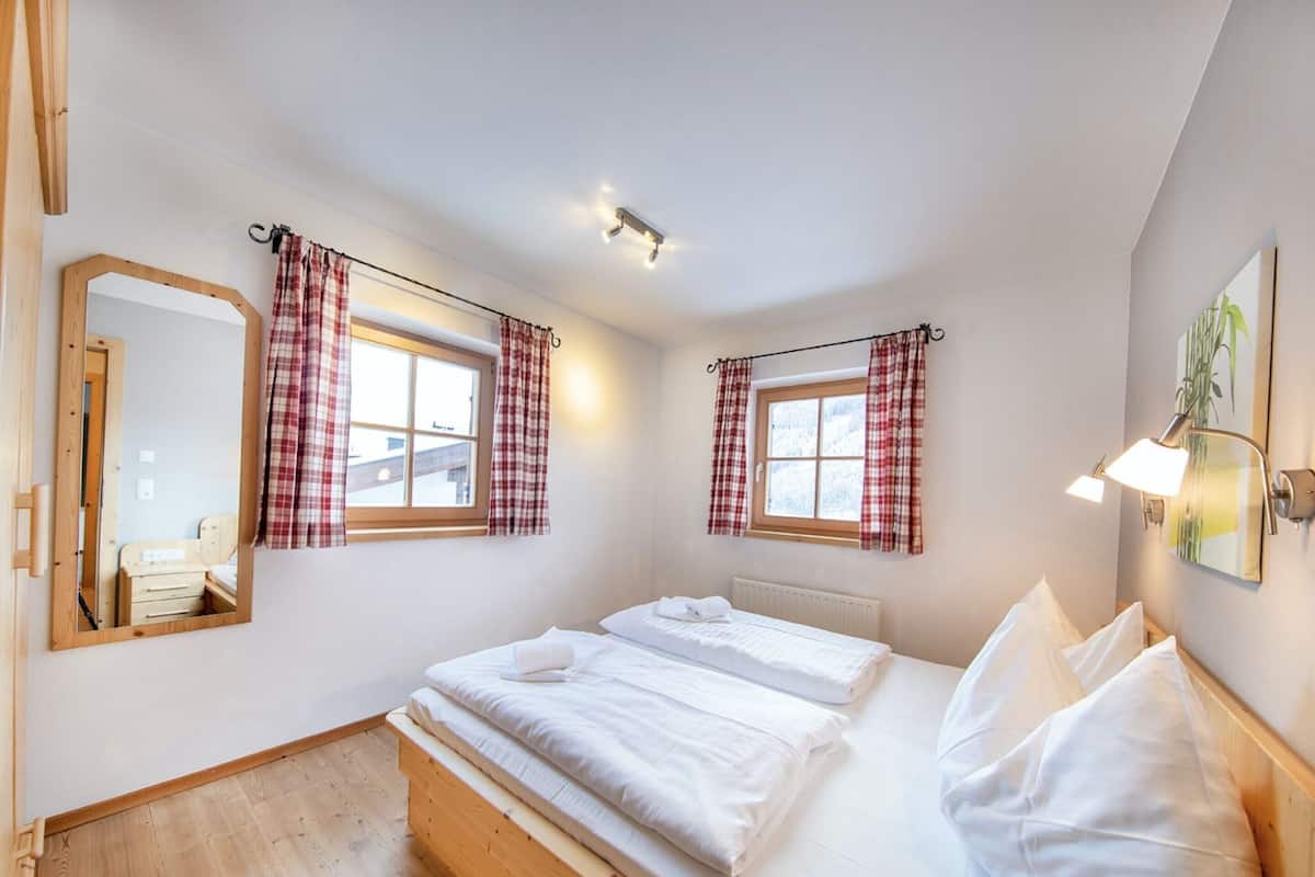 A bedroom with two beds, a wooden floor, a window with plaid curtains, a mirror, and a wall-mounted lamp.