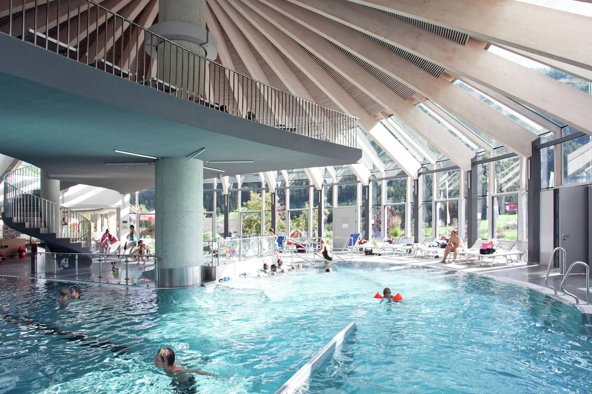 An indoor swimming pool with a modern architectural design, featuring a large central column and a sloped ceiling with skylights.
