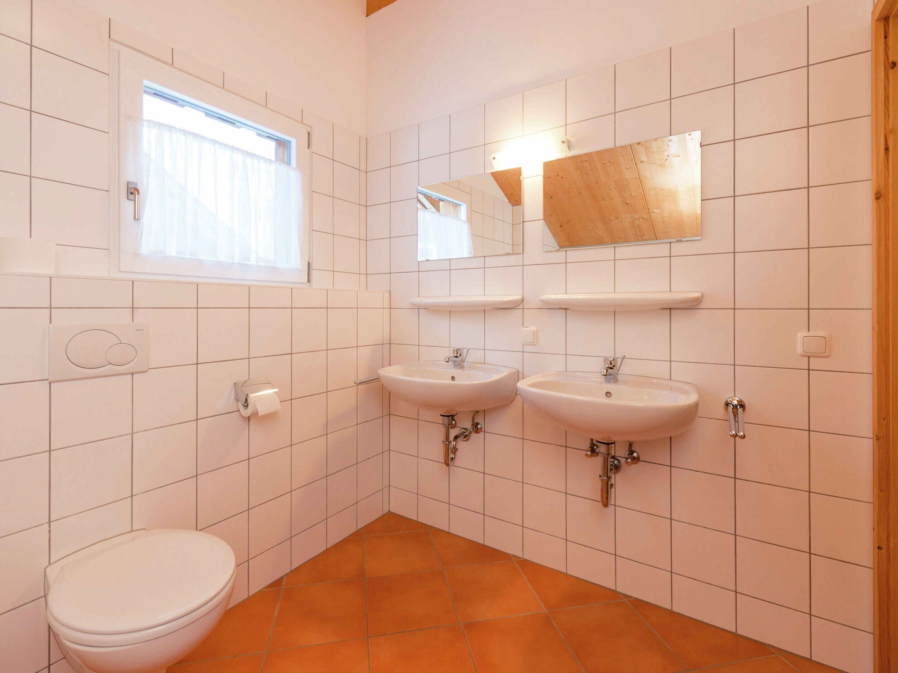 A bathroom with white tiled walls, two sinks, a toilet, a window with curtains, and a mirror with a wooden frame.