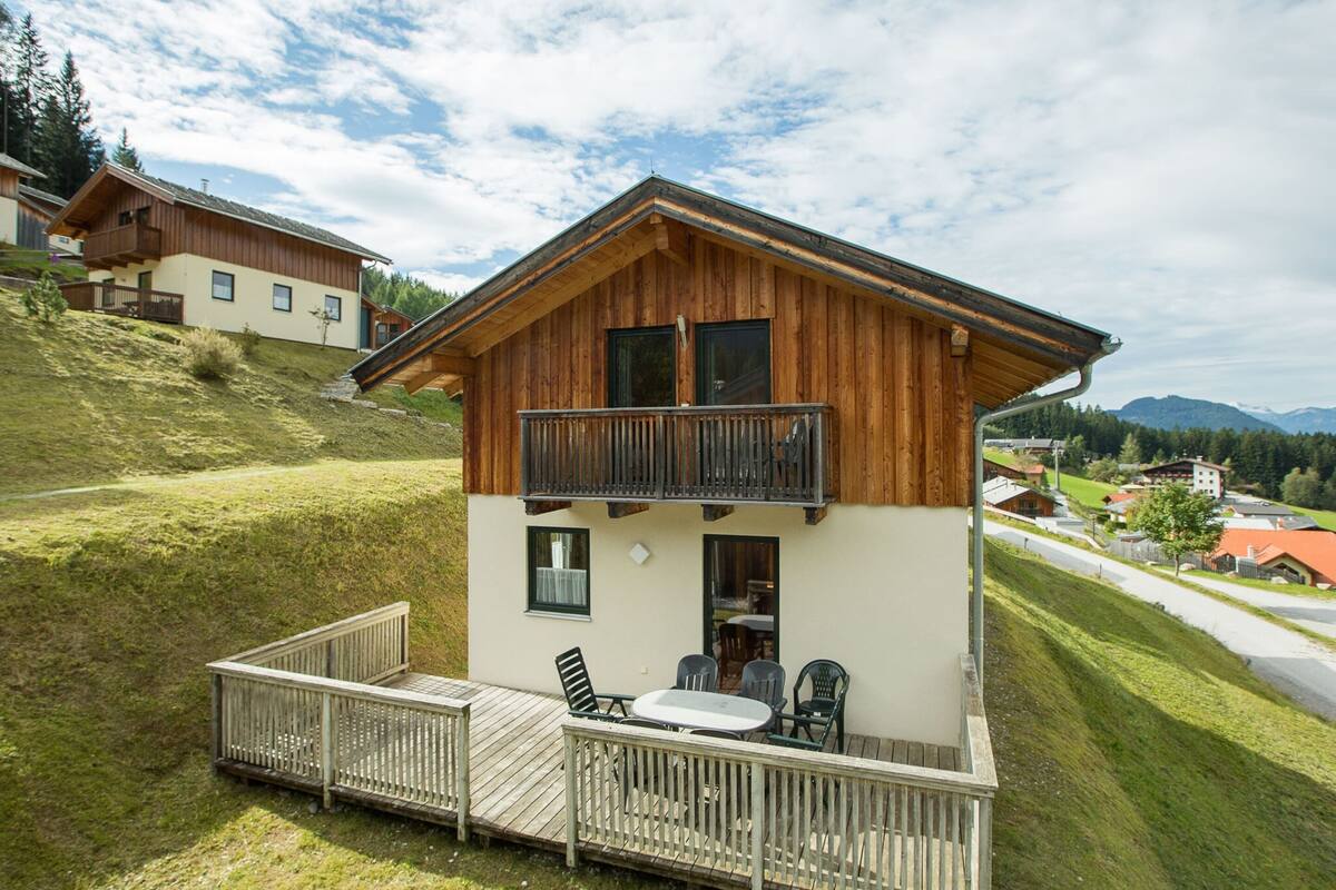 Un chalet de madera de dos pisos con balcón, una terraza con asientos exteriores y vista a un paisaje montañoso.