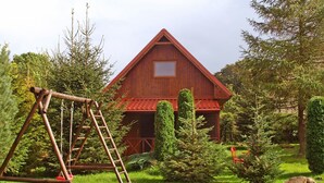 A rustic wooden barn with a red roof, surrounded by lush greenery and a swing set. - Authentic Holiday Home in Kopalino With Fireplace (Choczewo)