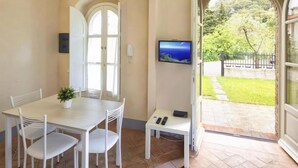A dining area with a white table and chairs, a small potted plant, a television on the wall, and a view of a garden through an open door. - Apartment in Lerici With Garden (Lerici)