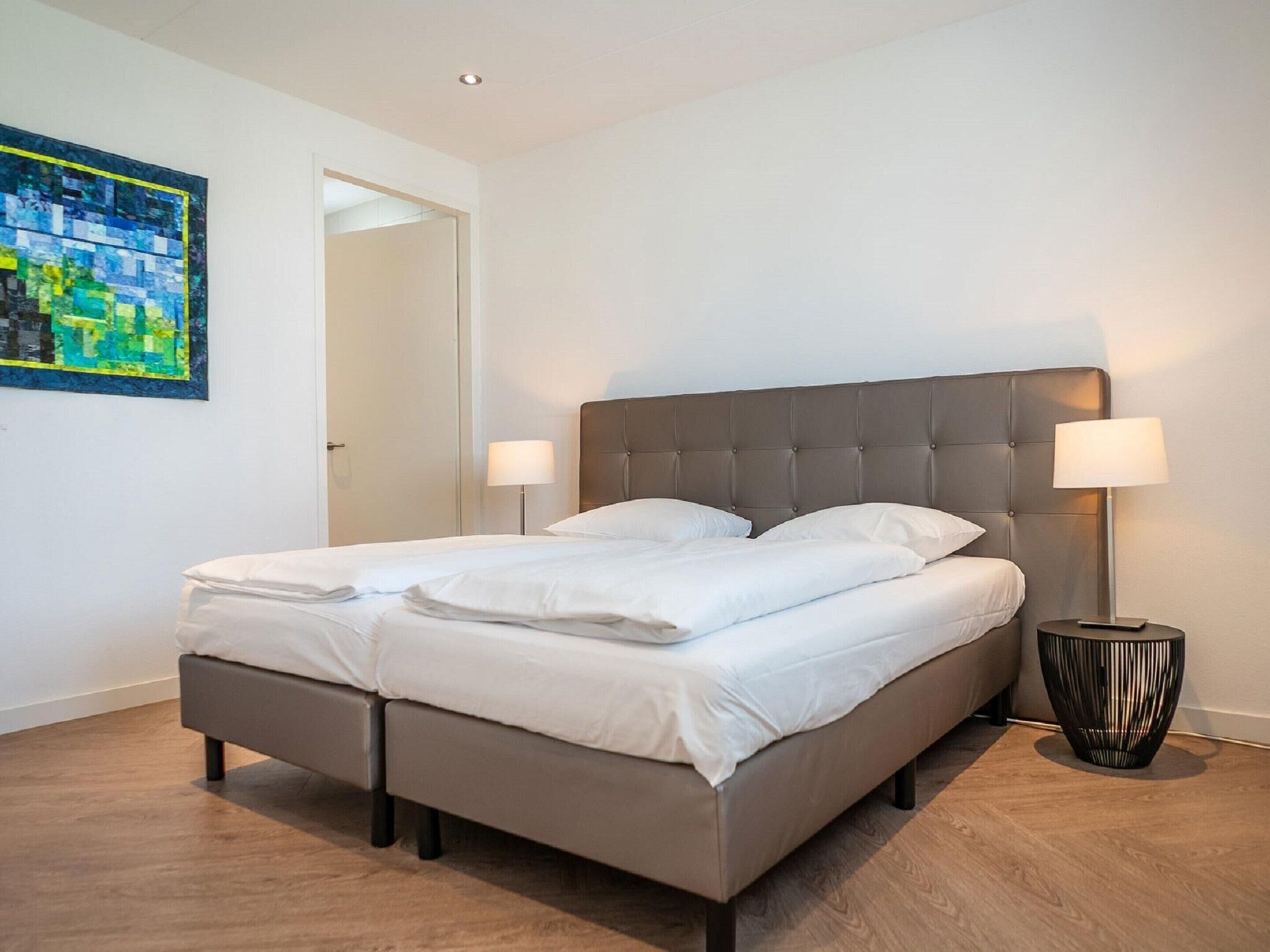 Une chambre moderne avec un lit recouvert d’un tissu gris, deux lampes de chevet et un plancher en bois.