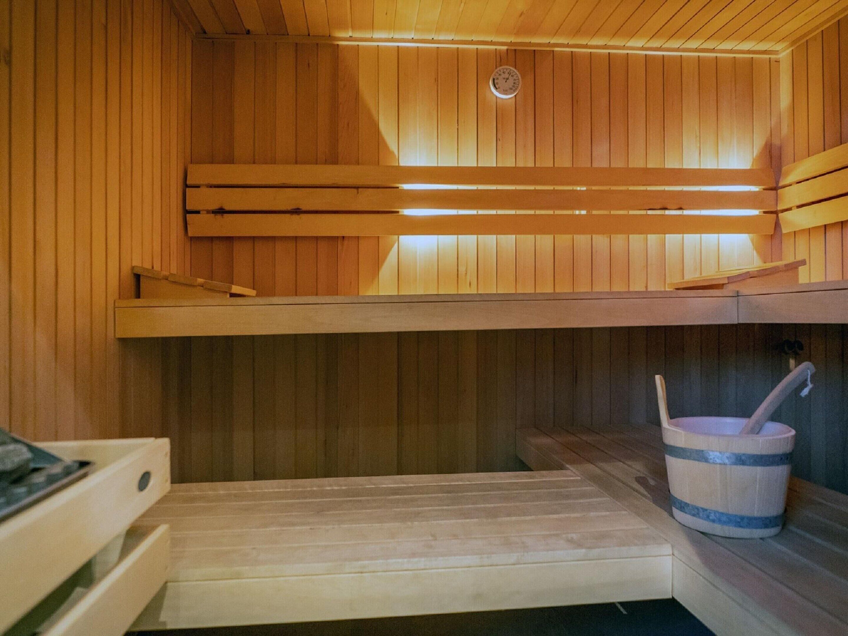 Una sauna con paredes y bancos de madera, techo de madera con un reloj, y un cubo con un cepillo.