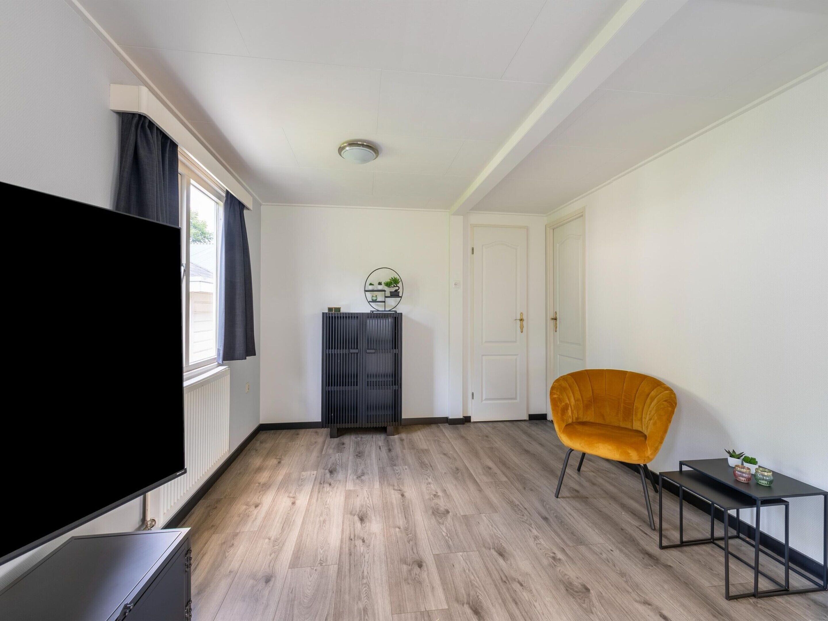 A modern living room with a flat-screen TV, a wooden floor, a black entertainment unit, a yellow armchair, and a small side table.