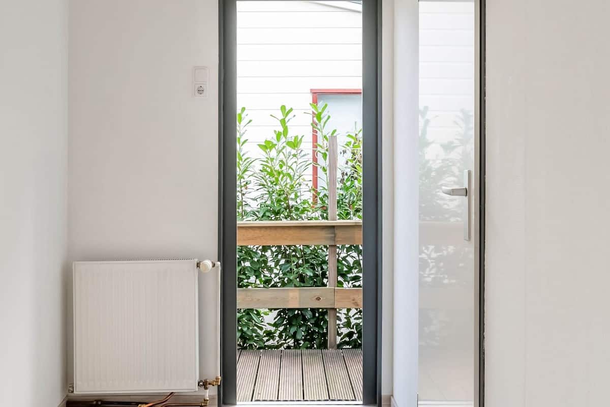 Une pièce avec un radiateur, une porte donnant sur un balcon agrémenté de plantes et une fenêtre avec des stores.