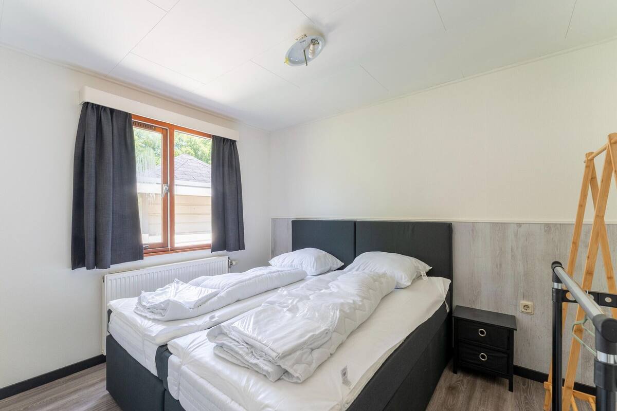 Un lit double avec du linge de lit blanc, une fenêtre avec des rideaux foncés, un radiateur, une petite table de chevet avec des tiroirs et une échelle en bois.