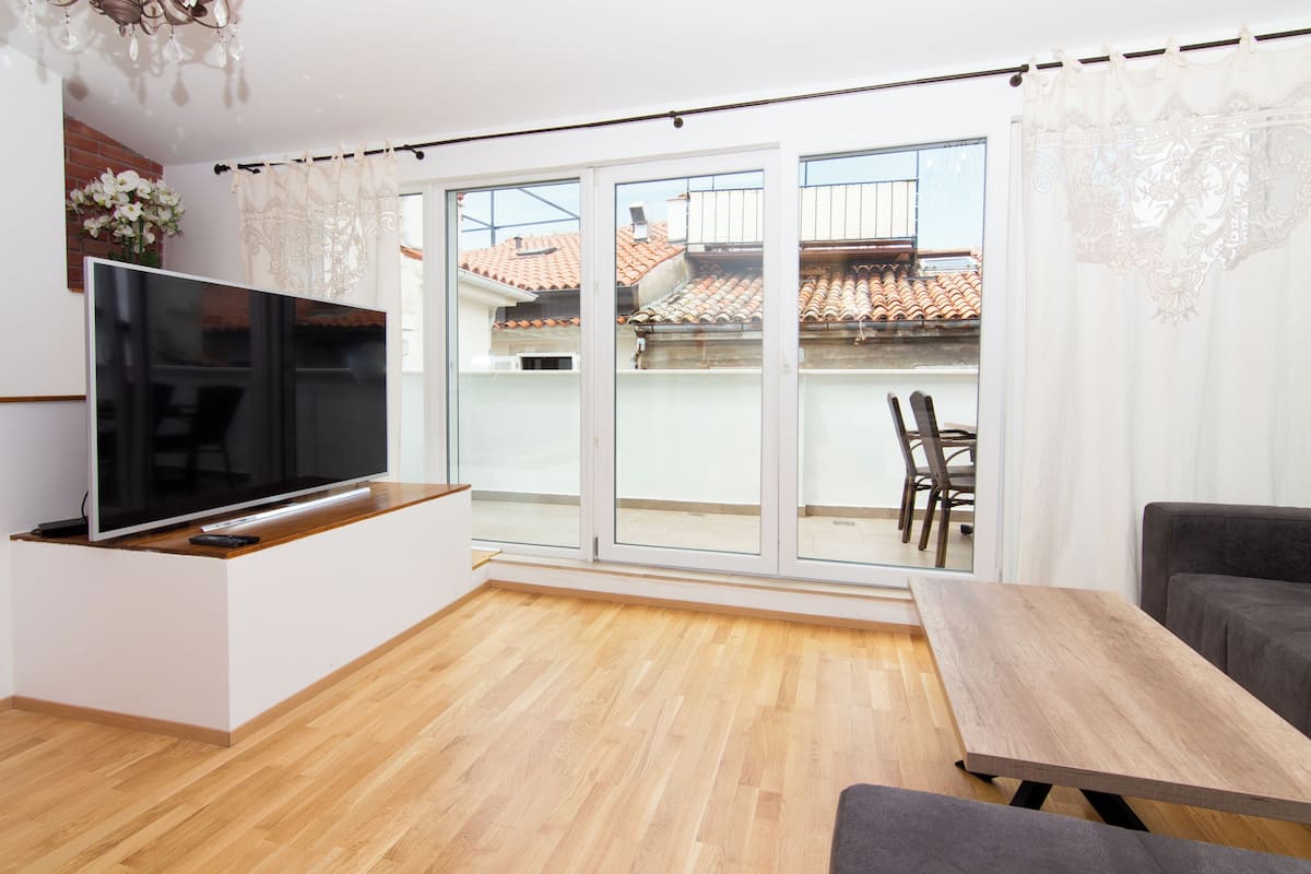 A modern living room with a flat-screen TV, wooden flooring, a grey sofa, and a dining table.