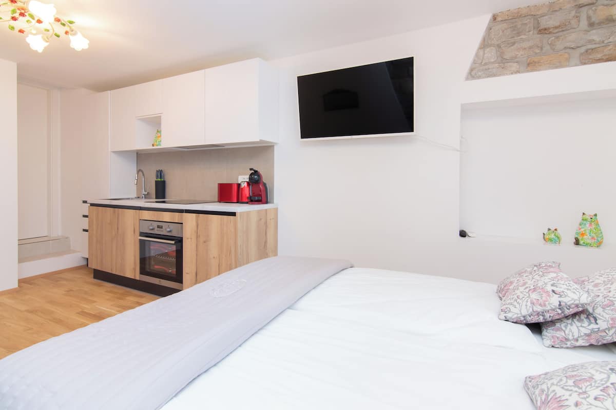 A modern bedroom with a bed, a small kitchenette, a TV mounted on the wall, and a stone accent wall.