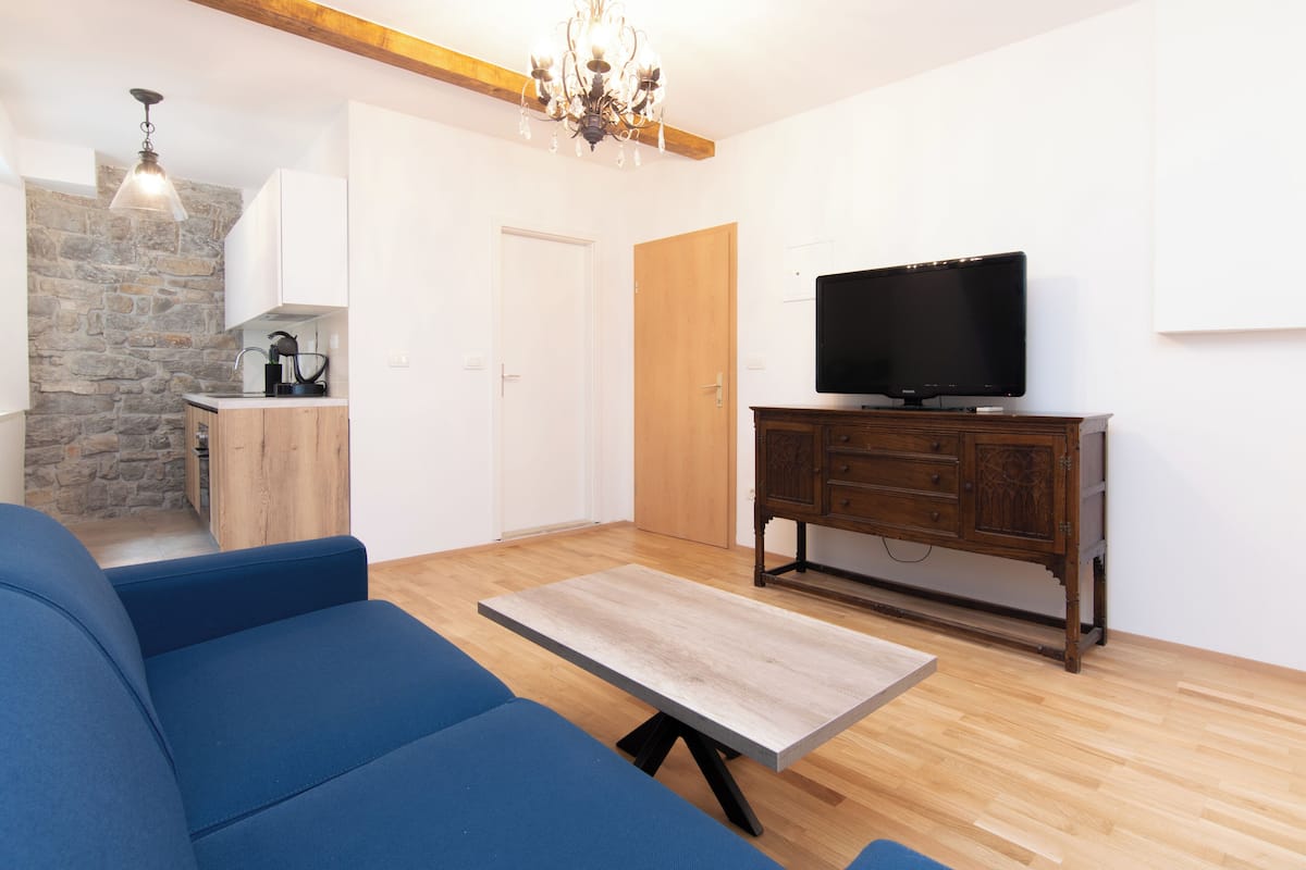 A living room with a blue sofa, a wooden coffee table, a TV on a wooden cabinet, a chandelier, and a stone wall.