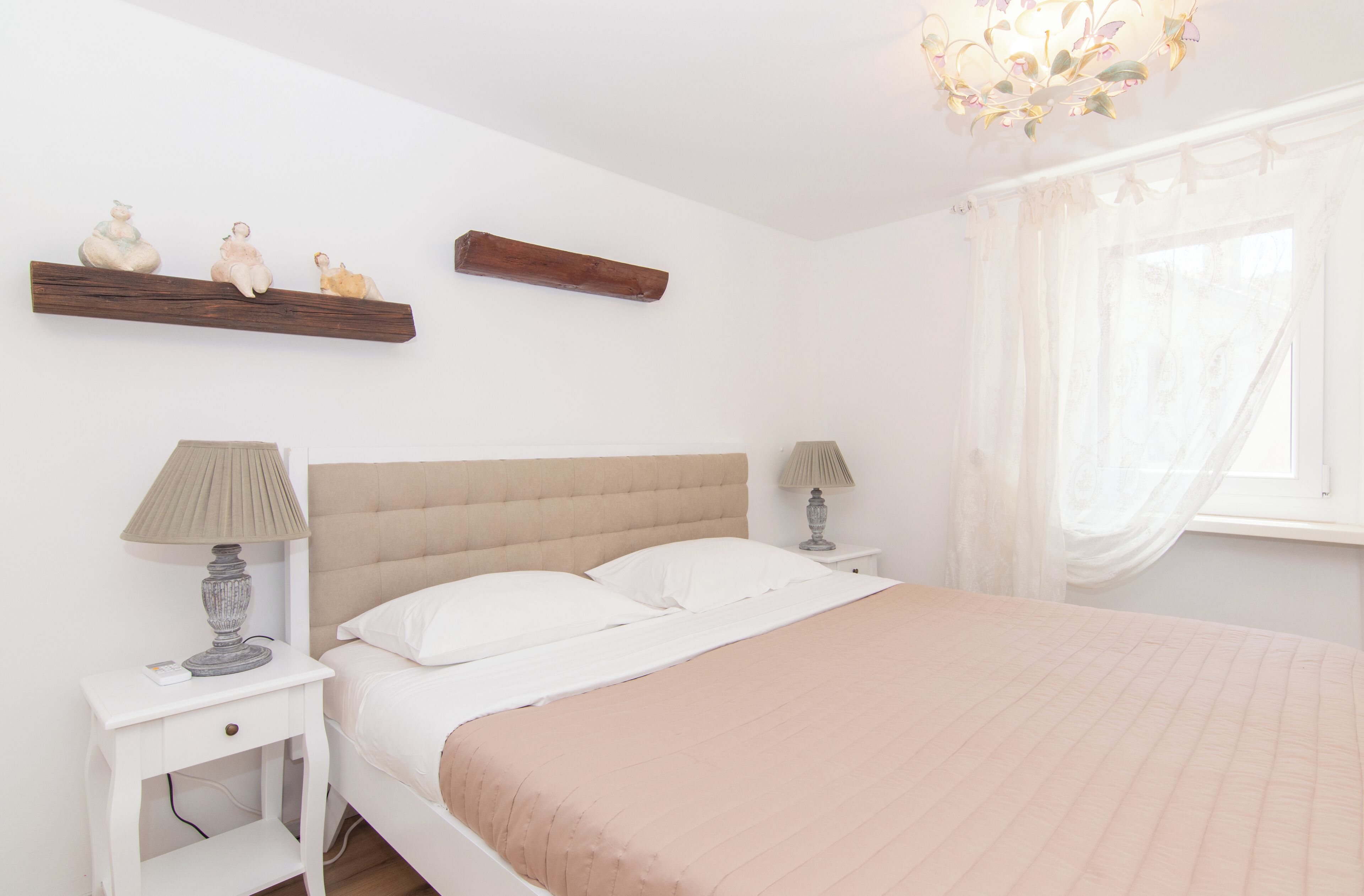 Een slaapkamer met een bed, nachtkastjes, een lamp, een plank met beeldjes, een raam met dunne gordijnen en een houten balk boven de plank.