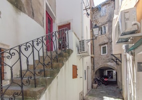 A narrow alleyway with a staircase leading to a building with a red door. There is a bicycle parked in the alley, and the walls are made of stone.