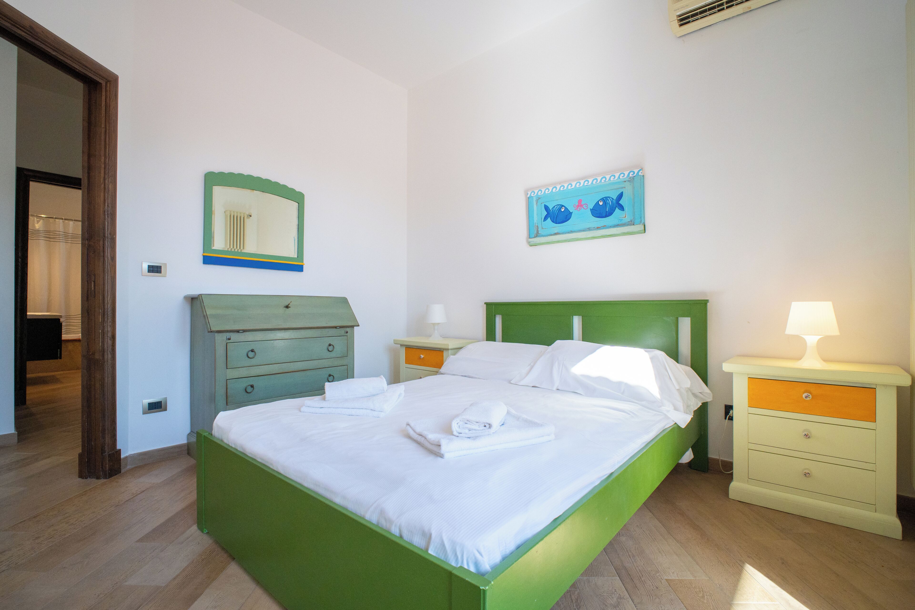 Ein Schlafzimmer mit einem grünen Bett, weißen Bettwäsche, einem Nachttisch mit Lampe, einer Kommode, einem Spiegel und einem Wandbild.