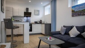 A modern kitchen with white cabinets, a central island, and a dining area with a bar stool. - HANNIBAL - Design Apartment Lavendel 4 (Wolfsburg)