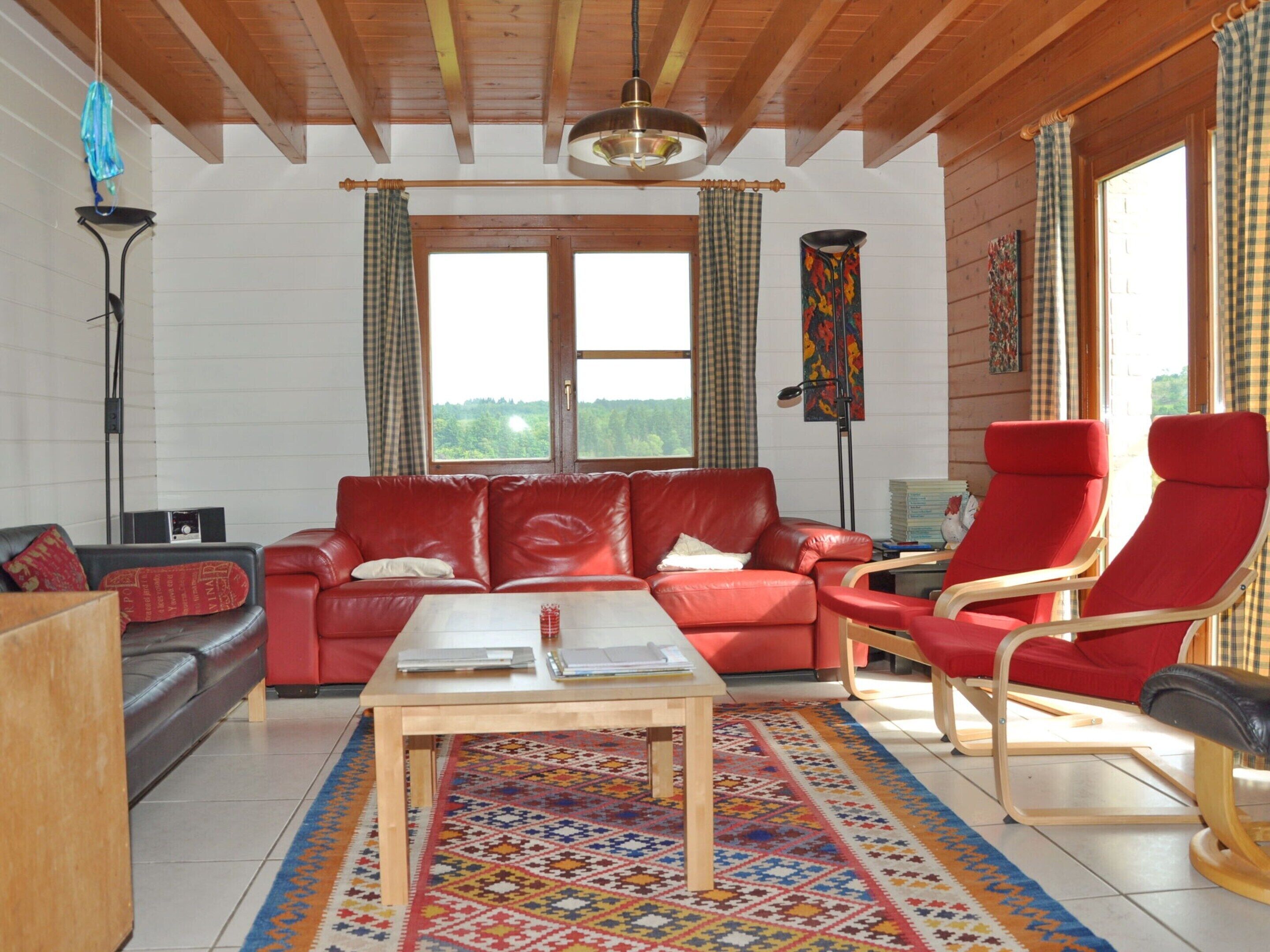 A living room with a red leather sofa, a wooden coffee table, and red armchairs. There are two windows with curtains, a floor lamp, and a colorful rug.