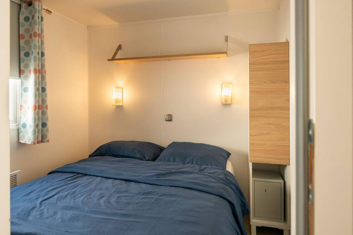 Un dormitorio con una cama, dos luces fijadas en la pared, un estante de madera y una cortina.
