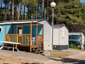 En blå campingvogn med en træterrasse og et picnicbord, omgivet af træer.