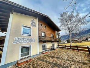 A traditional chalet with a wooden balcony, a stone-tiled patio, and a mountain backdrop.