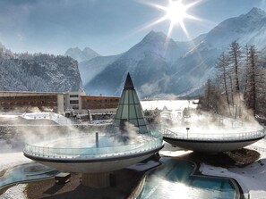Un área de spa con una fuente central, rodeada de montañas nevadas y un edificio con numerosas ventanas.