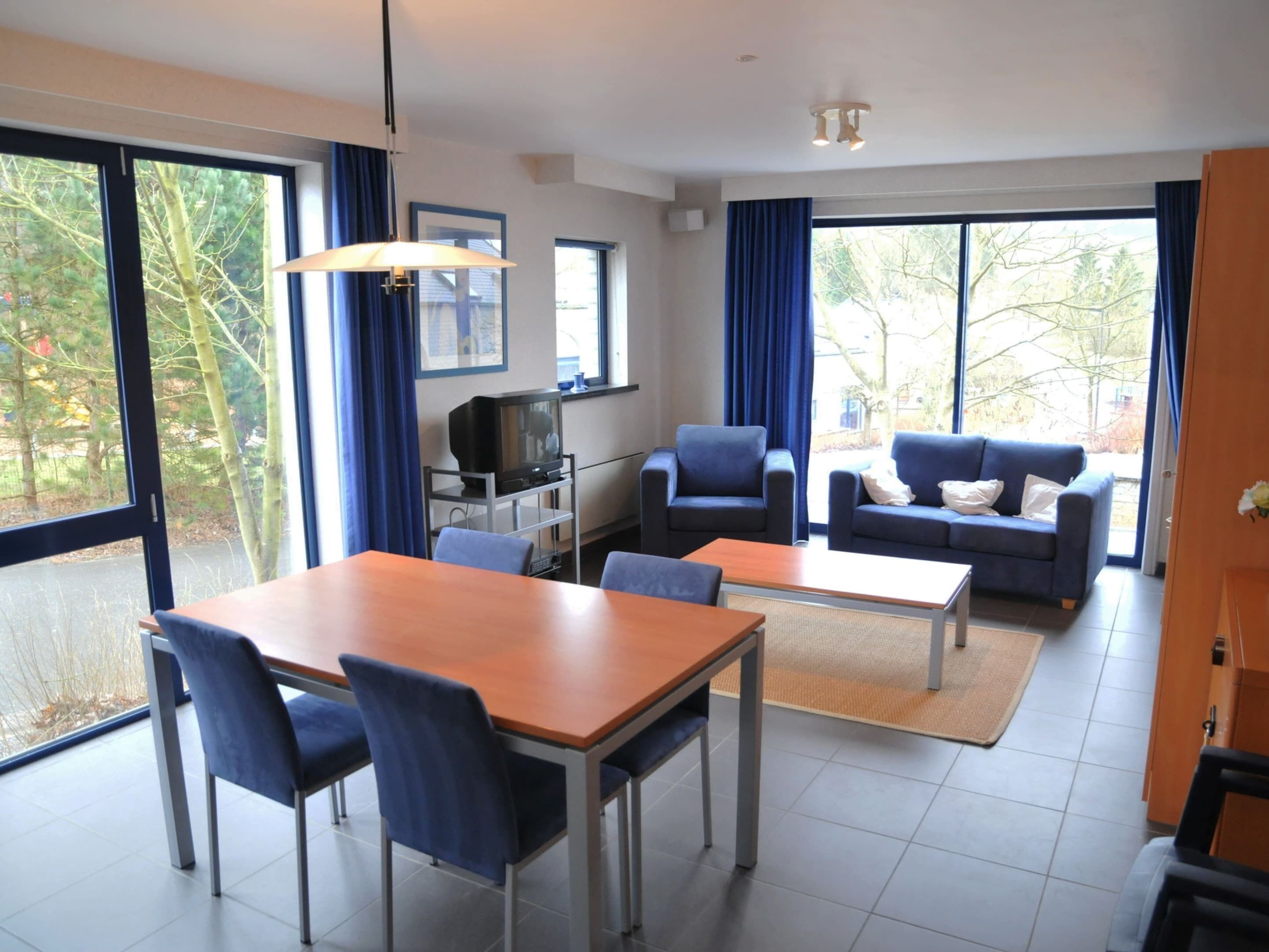A modern living room with a dining area, featuring a TV, blue furniture, and large windows offering a view of trees.