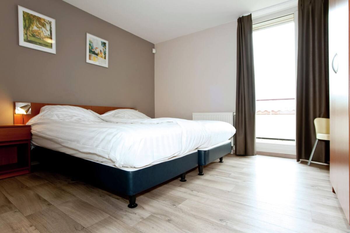 Une chambre à coucher comprenant un lit, une table de chevet, une fenêtre avec des rideaux et deux tableaux accrochés au mur.