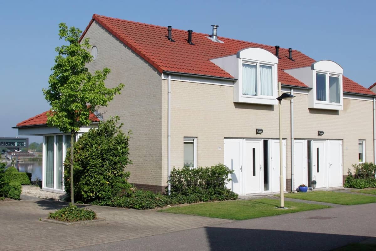 Un immeuble résidentiel de deux étages, avec un toit recouvert de tuiles rouges, un petit balcon et une fenêtre ornée d’un rideau blanc.